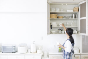 woman in white bright kitchen putting away white dishes in cabinet beside glasses and baskets