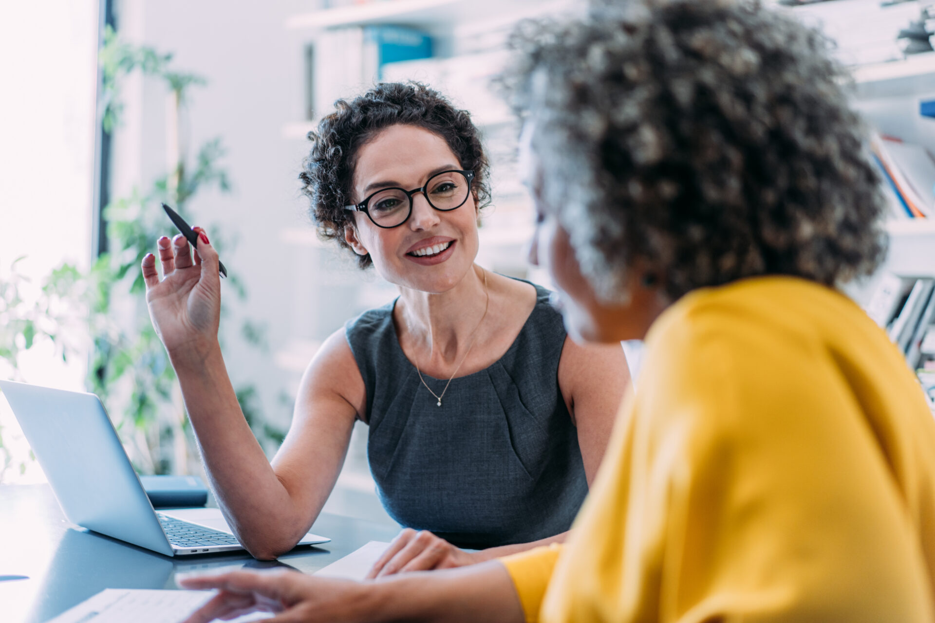 Professional woman in glasses discussing documents with a client in modern office setting with laptop and bookshelf.