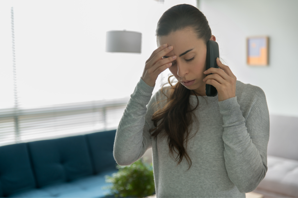 A worried woman in a gray sweater holding a phone to her ear while touching her forehead with her other hand. She appears stressed or concerned during a phone conversation in a modern living room with a blue couch and a window with blinds in the background.
