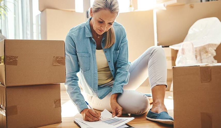 woman sitting in living room surrounded by moving boxes looking at checklist with pen in hand