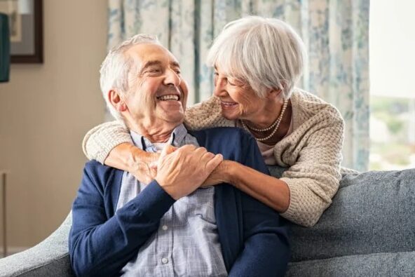 Happy elderly couple sitting on a couch in a cozy living room. The woman embraces the man from behind, both smiling warmly at each other. The room features soft furnishings, framed artwork, and natural light coming through floral curtains.